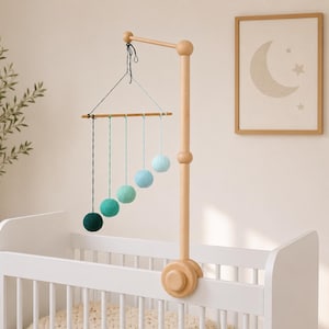 Puede incluir: Un móvil de madera para cuna con un brazo de soporte vertical sujeto a la estructura. Cinco pompones de lana en un degradado de tonos verdes y azules cuelgan de una barra horizontal de madera mediante hilos finos, formando una línea diagonal.