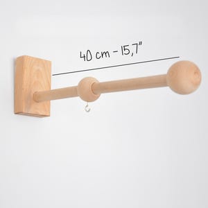 May include: A wooden curtain rod, 40 cm long, fixed to a wall. It features a rectangular base and a rounded end. The rod is made of light-coloured wood and includes a small metal hook. The text on the image reads "40 cm - 15.7".