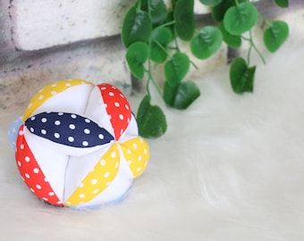 May include: A multi-colored fabric ball with white, red, yellow, blue, and navy blue polka dots. The ball is made of fabric squares sewn together to create a sphere.