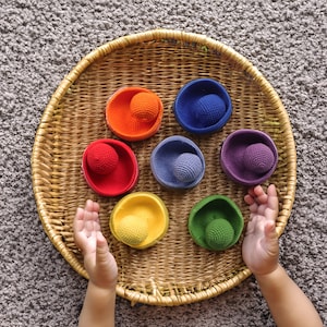 Puede incluir: Una bandeja de mimbre con siete bolas de crochet de colores vivos y cuencos a juego. Las bolas son roja, naranja, amarilla, verde, azul, morada e índigo.
