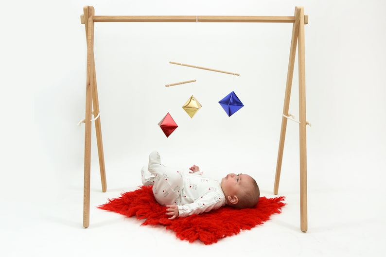 May include: A baby lying on a red fuzzy rug looking up at a wooden mobile with three diamond-shaped ornaments. The ornaments are red, gold, and blue.