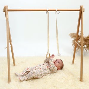 May include: A wooden baby play gym with two hanging toys, a wooden ring and a bell. A baby is lying on a white fluffy rug, looking up at the toys.