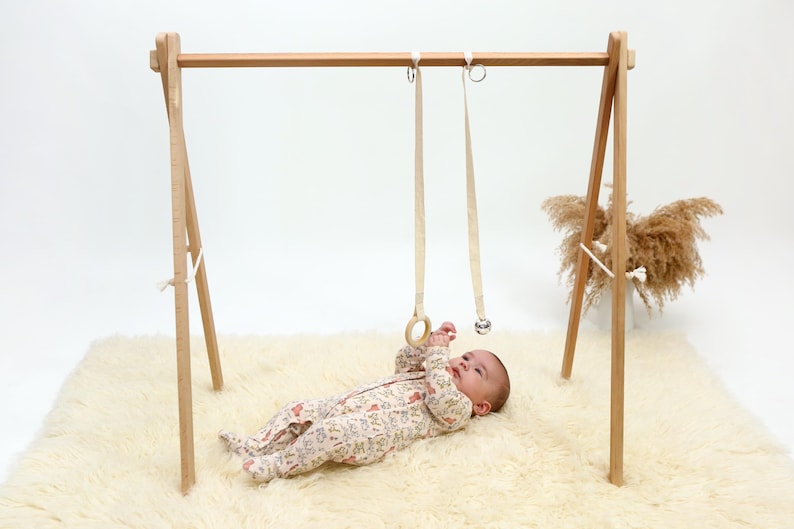 May include: A wooden baby play gym with two hanging toys, a wooden ring and a bell. A baby is lying on a white fluffy rug, looking up at the toys.