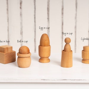 Peut inclure: Ensemble de jouets éducatifs en bois comprenant un cube dans une boîte, une boule dans une tasse, un œuf dans une tasse, une cheville dans une tasse et un cylindre dans une tasse. Chaque jouet est nommé.