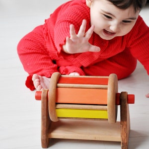 Può includere: Un giocattolo di legno con un cilindro rotante che presenta strisce colorate in rosso, arancione, giallo e verde. Il giocattolo è su una superficie bianca e un bambino gli sta tendendo la mano.