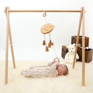 May include: A wooden baby play gym with a hanging wooden toy with bells and a ring. A baby is lying on a white fluffy rug, looking up at the toy.
