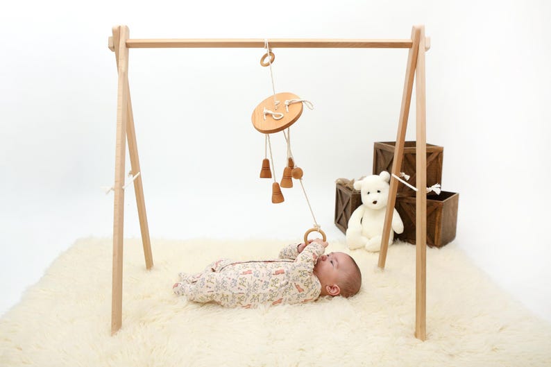 May include: A wooden baby play gym with a hanging wooden toy with bells and a ring. A baby is lying on a white fluffy rug, looking up at the toy.