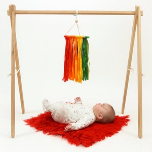 May include: A wooden baby play gym with a rainbow-colored tassel hanging from the top bar. A baby is lying on a red fuzzy rug looking up at the tassel.