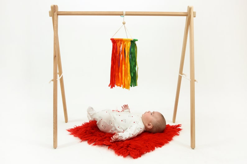 May include: A wooden baby play gym with a rainbow-colored tassel hanging from the top bar. A baby is lying on a red fuzzy rug looking up at the tassel.