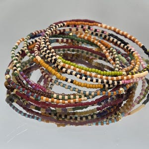 May include: A collection of colorful beaded bracelets in various designs. The bracelets feature small, round beads in shades of brown, orange, green, black, and white. Some bracelets have gold-colored accents. The bracelets are stacked and reflected on a shiny surface.