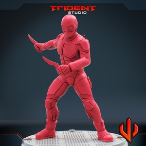 Pu&ograve; includere: Una action figure rossa di una persona che indossa una tuta intera e un casco, tenendo due coltelli in una posa di combattimento. La figura &egrave; in piedi su una base circolare con uno sfondo nero.