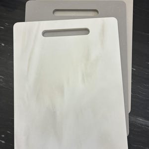 May include: Three rectangular cutting boards in shades of white, gray, and beige. Each board has a rectangular handle cut out at the top. The boards are stacked, with the white board in front.