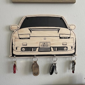 May include: A light-colored wooden wall-mounted key holder shaped like the front of a Nissan 180SX car. The design features black engraved lines detailing the headlights, grille, and bumper, with a license plate reading 180SX. Four metal hooks hold keys.