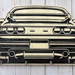 May include: A wooden key holder shaped like the rear of a car, featuring a black and natural wood color scheme. The design includes key hooks at the bottom and is mounted on a light-colored wooden surface.