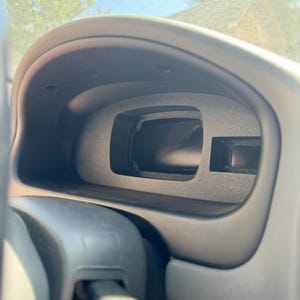 May include: A close-up of a car dashboard, featuring a black instrument cluster with various openings and a steering column. The dashboard has a matte finish and is angled towards the viewer, with a glimpse of the windshield and sky visible.