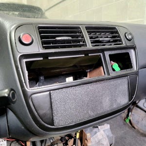 May include: A black car dashboard with a red button, two air vents, and a black plastic panel with a textured surface.