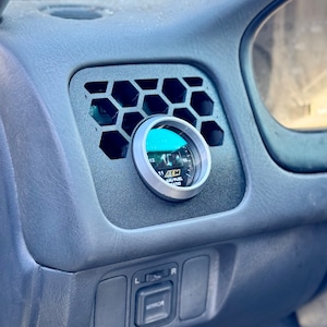 Può includere: Un cruscotto di un'auto nero e grigio con un indicatore rotondo che indica "AEM AIR/FUEL RATIO". Il misuratore è montato in una griglia di ventilazione esagonale.