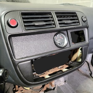 May include: A black car dashboard with a black and gray textured panel, a black and silver volt gauge, and a black rectangular cutout. The gauge reads "VOLT" and has numbers from 12 to 16.