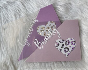 May include: Two purple and lavender coloured envelopes with heart-shaped leopard print accents. The word "Groceries" is written on the clear overlay of one envelope, and the word "Beauty" is written on the other.