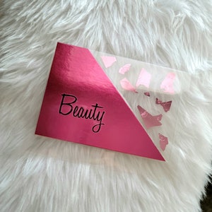 May include: A pink makeup palette with the word "Beauty" in cursive script. The palette has a shiny, metallic finish on the left side and a clear section with pink foil accents on the right. The palette rests on a fluffy white surface.