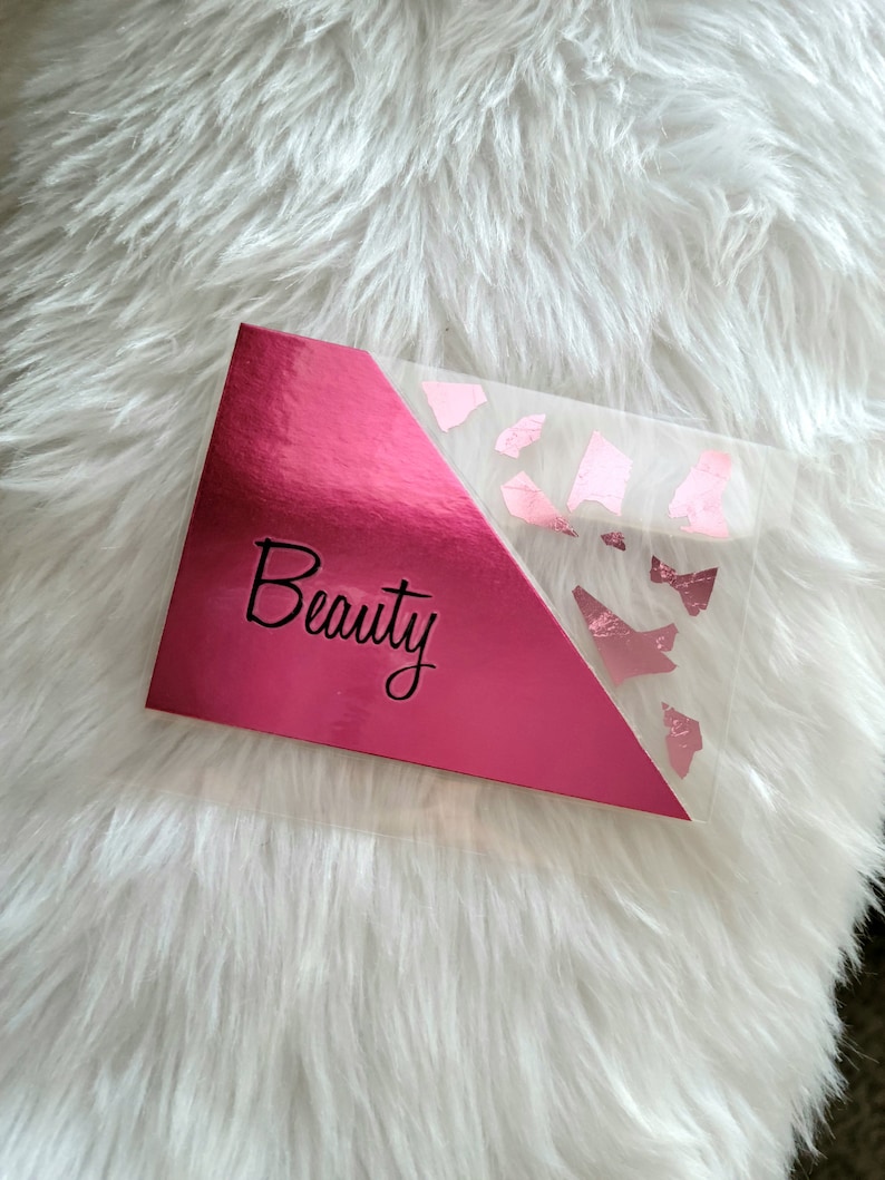 May include: A pink makeup palette with the word "Beauty" in cursive script. The palette has a shiny, metallic finish on the left side and a clear section with pink foil accents on the right. The palette rests on a fluffy white surface.