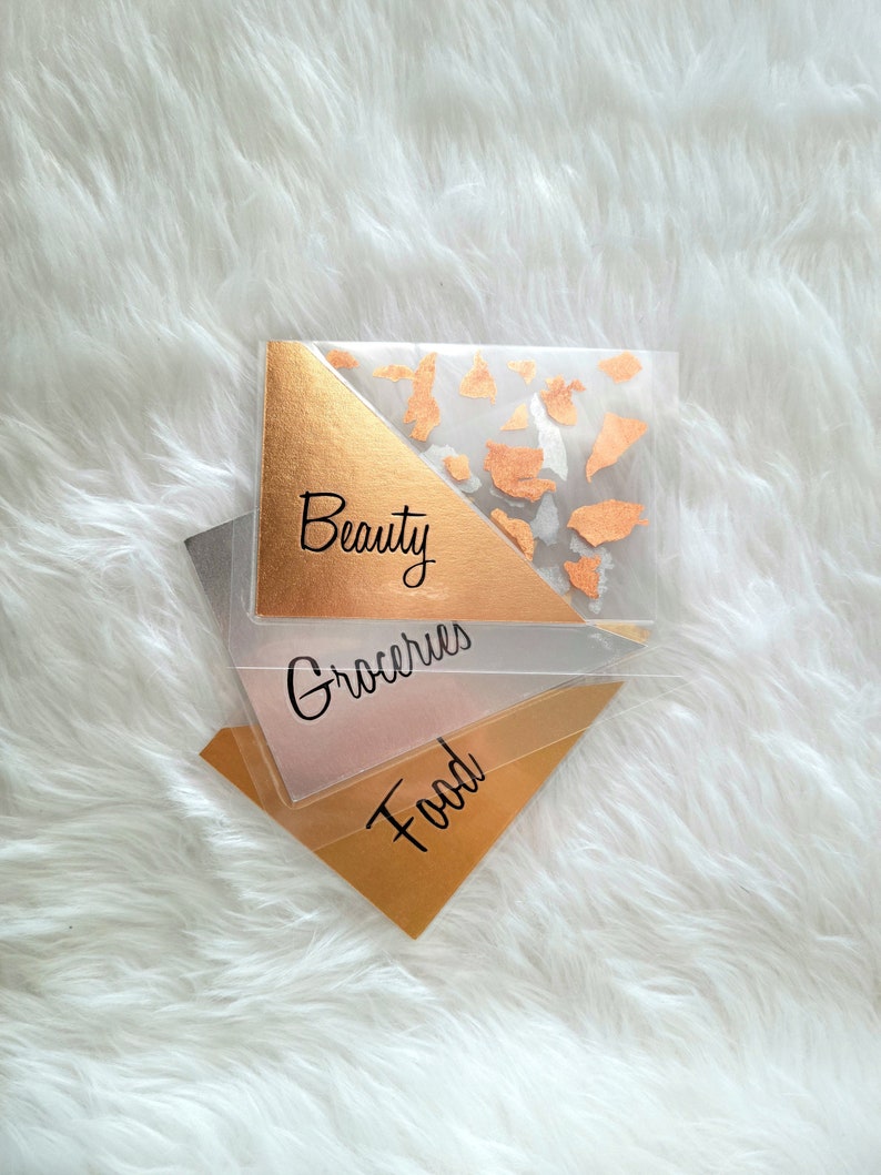 May include: Clear plastic budget envelopes with gold foil accents and black lettering. The envelopes are labeled "Beauty", "Groceries", and "Food". The background is a white, fluffy surface.
