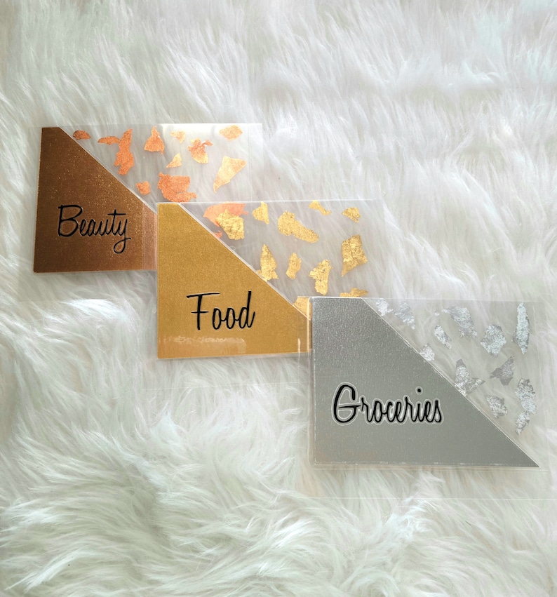 May include: Three clear acrylic budget organizers with metallic accents. Each organizer has a diagonal section in gold, copper, or silver, with gold, copper, or silver foil flakes. The labels read "Beauty", "Food", and "Groceries".
