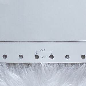 May include: A close-up of a white paper with several evenly spaced holes along the bottom edge. The text "A7" is written above a 2 cm measurement, indicating the distance between two holes. The paper has a folded top edge.