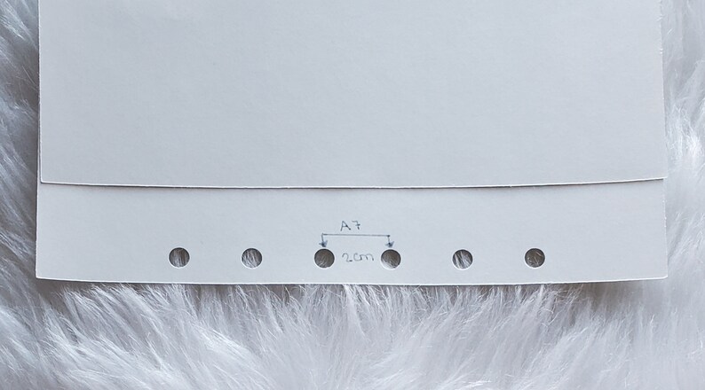 May include: A close-up of a white paper with several evenly spaced holes along the bottom edge. The text "A7" is written above a 2 cm measurement, indicating the distance between two holes. The paper has a folded top edge.