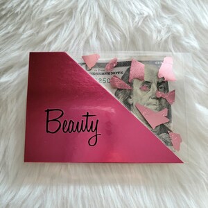 May include: A clear plastic wallet with a pink metallic accent and the word "Beauty" in black script. The wallet contains a US $100 bill with pink confetti.
