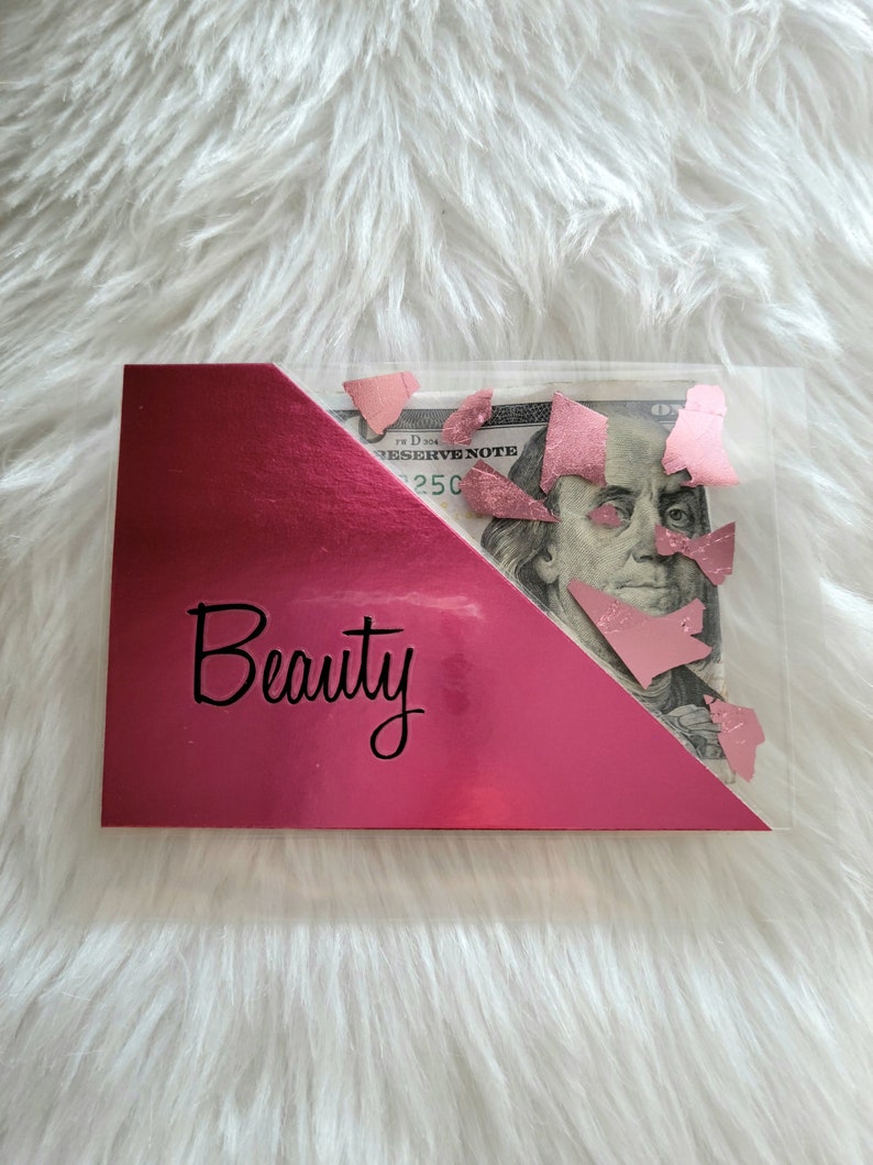 May include: A clear plastic wallet with a pink metallic accent and the word "Beauty" in black script. The wallet contains a US $100 bill with pink confetti.