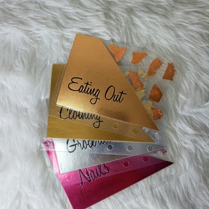 May include: A stack of budget envelopes in gold, silver, and pink. Each envelope is labeled with categories like "Eating Out," "Clothing," "Groceries," and "Nails." The gold envelope has orange foil accents.