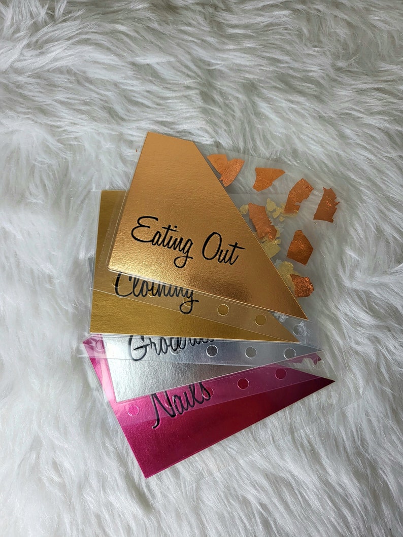 May include: A stack of budget envelopes in gold, silver, and pink. Each envelope is labeled with categories like "Eating Out," "Clothing," "Groceries," and "Nails." The gold envelope has orange foil accents.