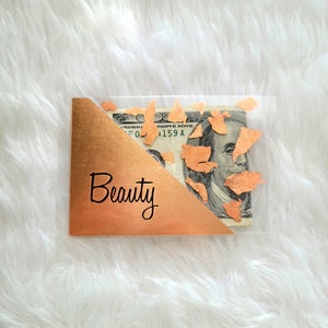 May include: A clear plastic gift card holder with a gold foil corner and the word "Beauty" in black script. A one hundred dollar bill is visible inside the holder, with gold confetti accents.