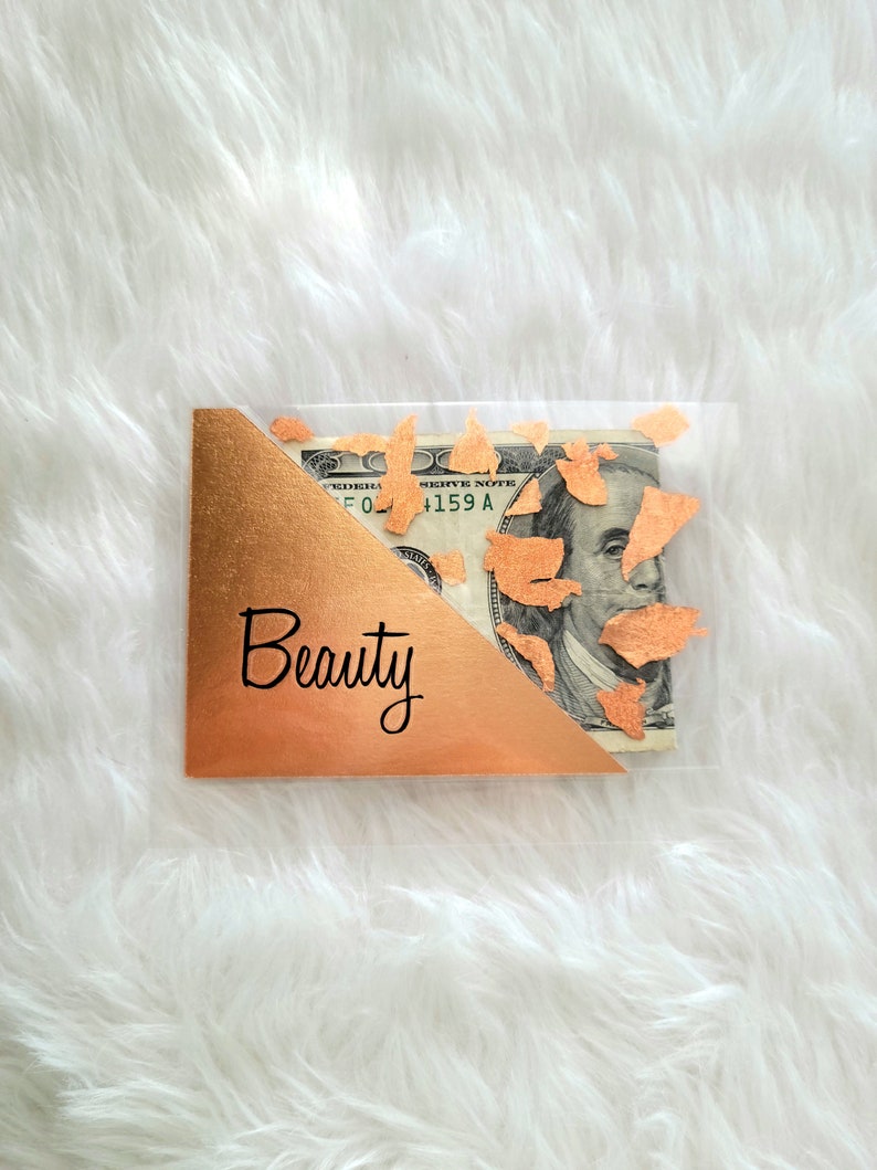 May include: A clear plastic gift card holder with a gold foil corner and the word "Beauty" in black script. A one hundred dollar bill is visible inside the holder, with gold confetti accents.