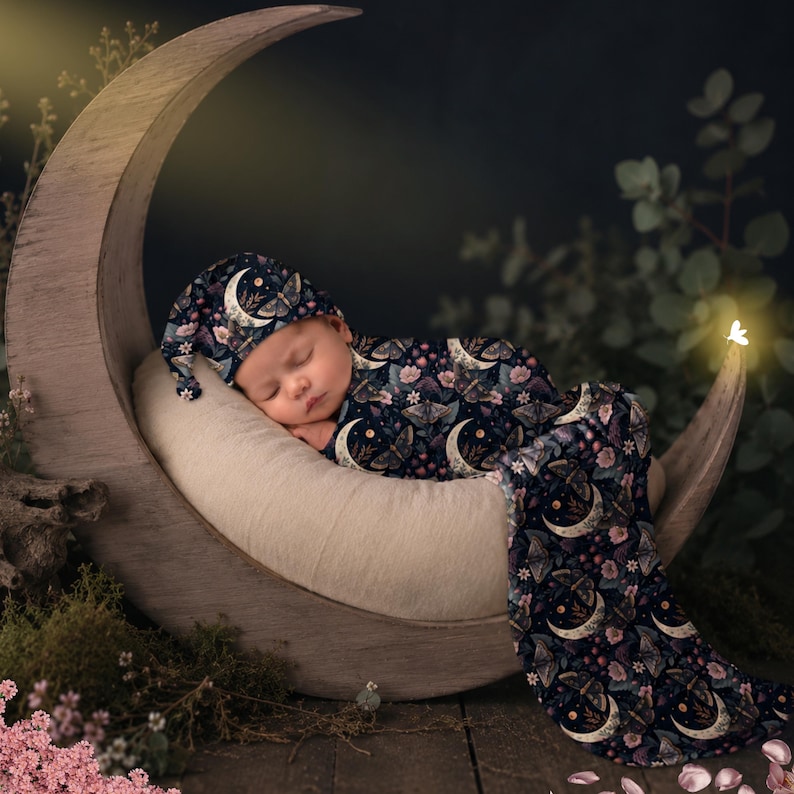 May include: A sleeping baby wearing a navy blue moon and moth-patterned sleep sack and hat, nestled in a wooden crescent moon. The sleep sack and hat feature a floral and crescent moon design. The scene is set against a dark background with foliage.