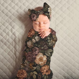 May include: A newborn baby wrapped in a floral swaddle blanket and matching headband. The dark background is covered with large, vintage-style flowers in shades of brown, cream, and purple. The baby is lying on a quilted, off-white surface.