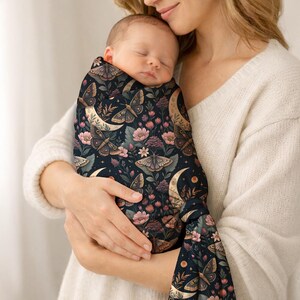 May include: A sleeping baby wrapped in a dark blue swaddle blanket. The blanket features a pattern of moths, crescent moons, and flowers in shades of brown, pink, and cream. The baby is held by a person wearing a white sweater.