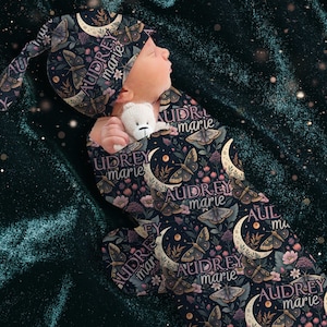 May include: A newborn baby swaddled in a dark blue, patterned sleep sack and matching cap. The pattern includes moths, moons, and floral elements. The name "AUDREY marie" is printed on the fabric. The baby is holding a small white teddy bear.