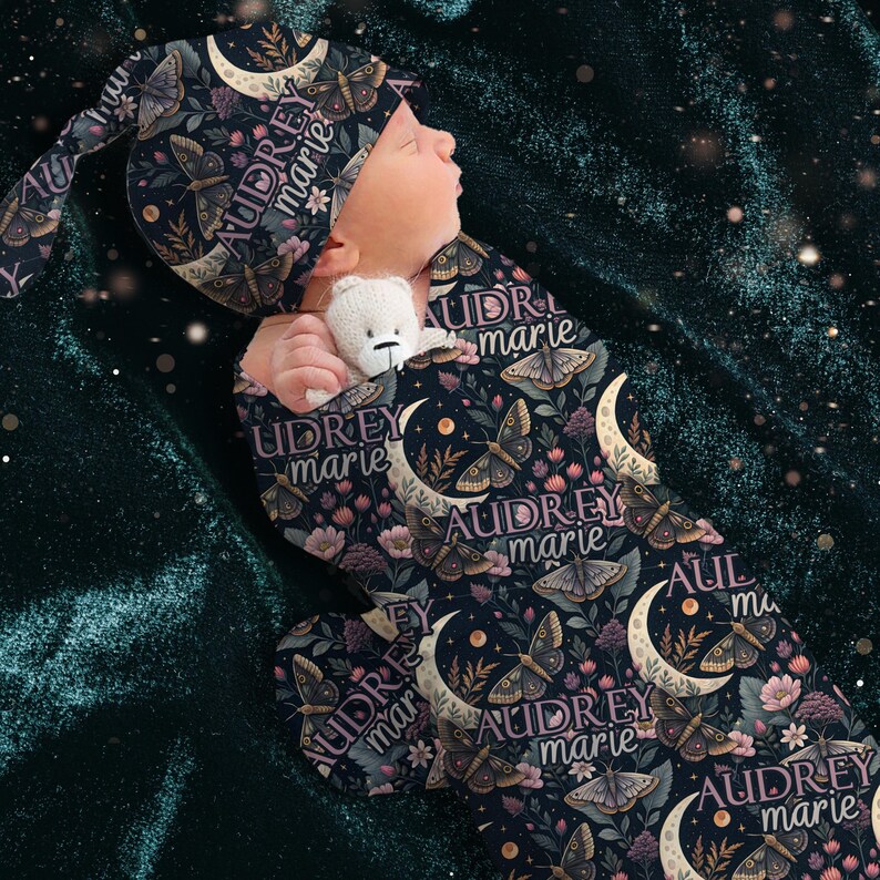 May include: A newborn baby swaddled in a dark blue, patterned sleep sack and matching cap. The pattern includes moths, moons, and floral elements. The name "AUDREY marie" is printed on the fabric. The baby is holding a small white teddy bear.