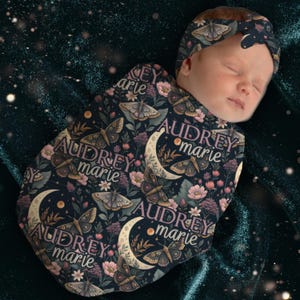 May include: A baby swaddled in a dark blue blanket and matching headband, both adorned with a moth and floral pattern. The name "Audrey Marie" is printed on the blanket. The baby is sleeping.