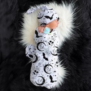 May include: A white swaddle with a black and white skull, crescent moon, bat, and star pattern. A matching black and white skull, crescent moon, bat, and star pattern hat is on the baby's head.