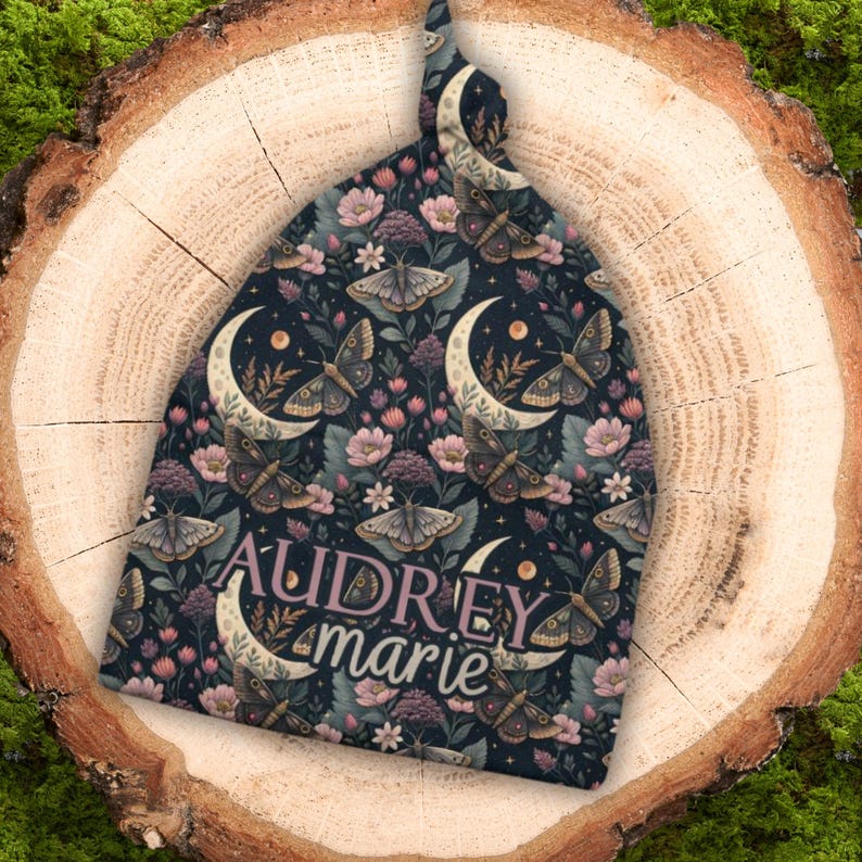 May include: A dark blue baby beanie with a repeating pattern of moths, crescent moons, and flowers. The name "Audrey Marie" is printed in a cursive font across the front of the hat. The hat is made of soft fabric.