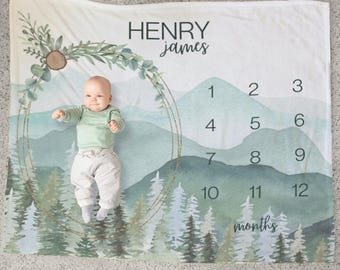 May include: A baby milestone blanket with a watercolor mountain and forest design. The blanket features a gold circle with greenery and a number chart for each month. The text "HENRY james" is at the top of the blanket.
