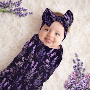 May include: A baby wrapped in a dark purple swaddle blanket and matching headband, both adorned with a lavender floral pattern. The baby is lying on a white, fluffy surface, with sprigs of lavender flowers around.