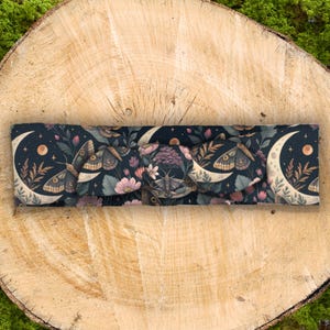 May include: A dark blue headband with a repeating pattern of moths, crescent moons, stars, and flowers. The headband is made of soft fabric and has a knotted design in the center. The color palette includes shades of blue, beige, pink, and purple.