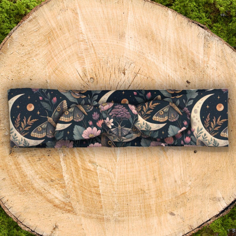 May include: A dark blue headband with a repeating pattern of moths, crescent moons, stars, and flowers. The headband is made of soft fabric and has a knotted design in the center. The color palette includes shades of blue, beige, pink, and purple.