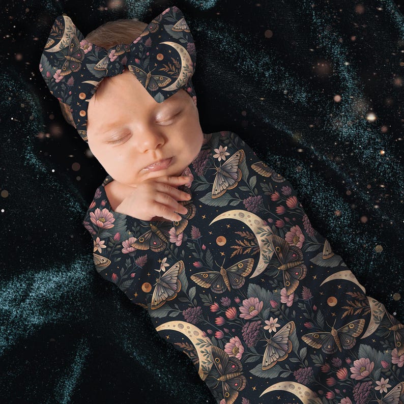 May include: A baby wearing a dark navy blue swaddle and matching headband, both adorned with a celestial pattern of moths, moons, and floral elements in shades of pink, beige, and green. The baby is sleeping peacefully.