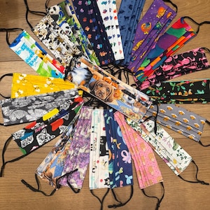 May include: A collection of assorted face masks with various patterns, including pandas, paw prints, dogs, and flamingos. Each mask has adjustable black ear loops. The masks are made of fabric and are arranged in a circular pattern.