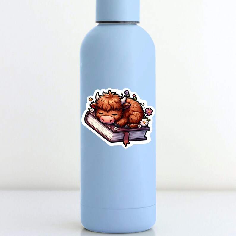 May include: A brown and white highland cow is sleeping on a closed book with a brown cover. The book is surrounded by small purple, pink, and white flowers.